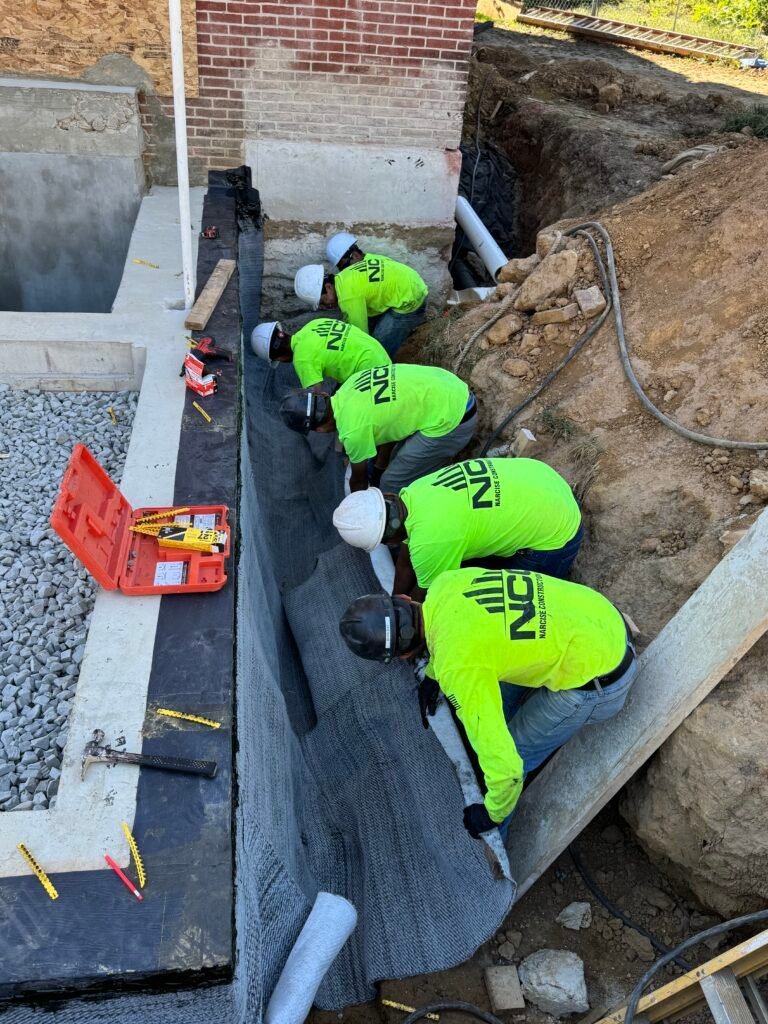 Four construction workers in neon shirts and hard hats install waterproofing membrane along a building foundation wall at an outdoor construction site. Tools and materials are visible nearby.