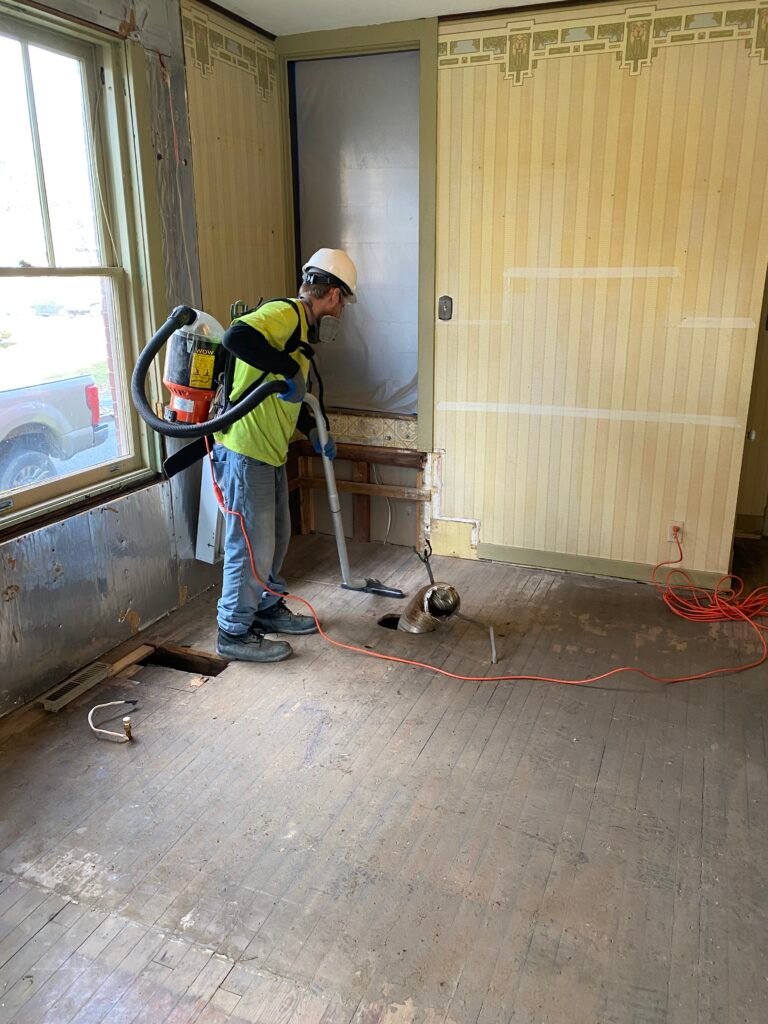 A worker wearing a hard hat and safety vest vacuums debris from a stripped wooden floor in a partially renovated room.
