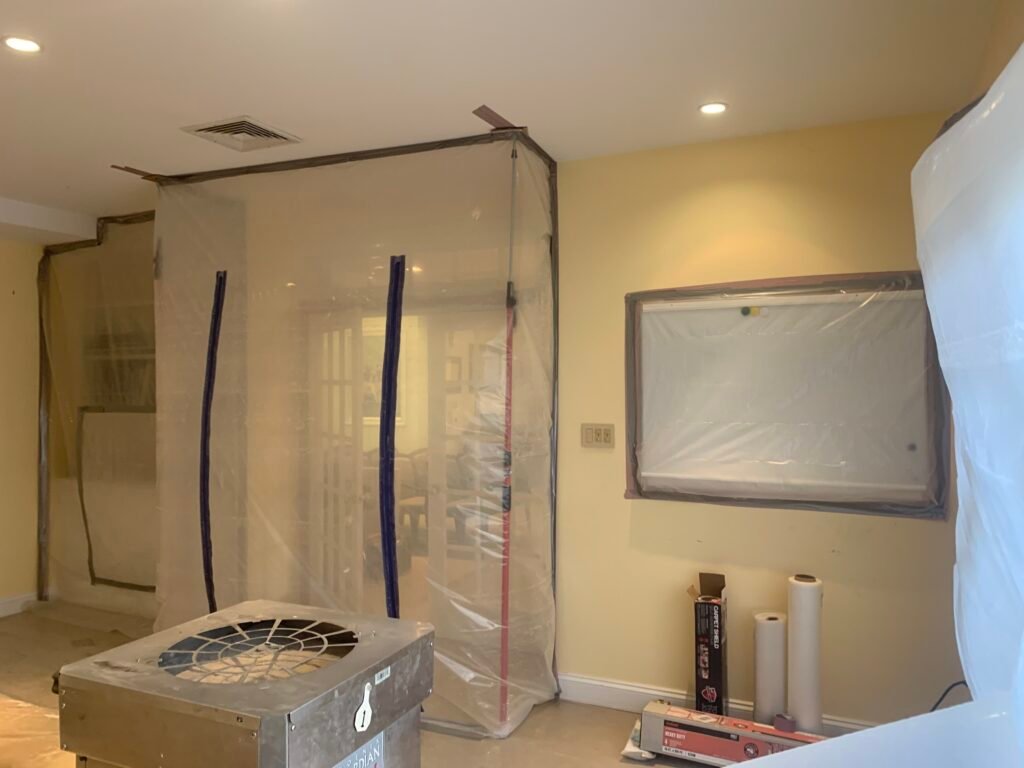 A room undergoing renovation with plastic sheeting covering doorways and a window, tools and materials on the floor, and yellow walls visible.