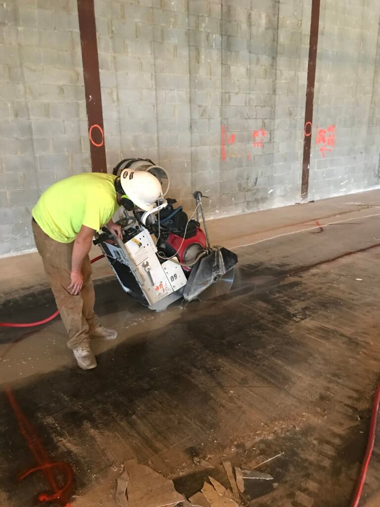 A construction worker in a yellow shirt operates a concrete saw, cutting through a floor inside a building with cinder block walls.