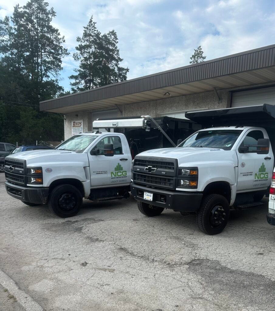 Two white NCG service trucks are parked outside a commercial building with trees and a cloudy sky in the background.