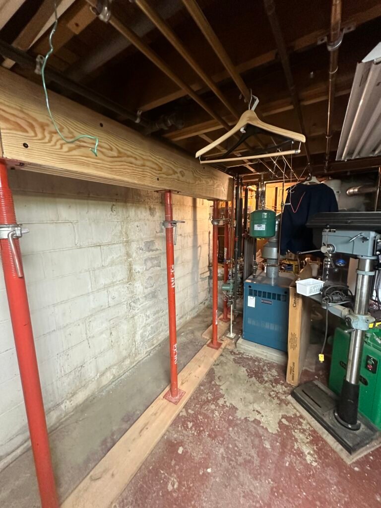 A basement with exposed ceiling beams, red support poles, a wooden beam, boiler equipment, a hanging clothes hanger, and various tools and boxes.