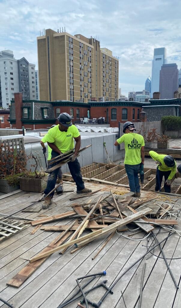 Three construction workers in neon shirts and helmets dismantle wooden decking on a rooftop, with city buildings visible in the background.