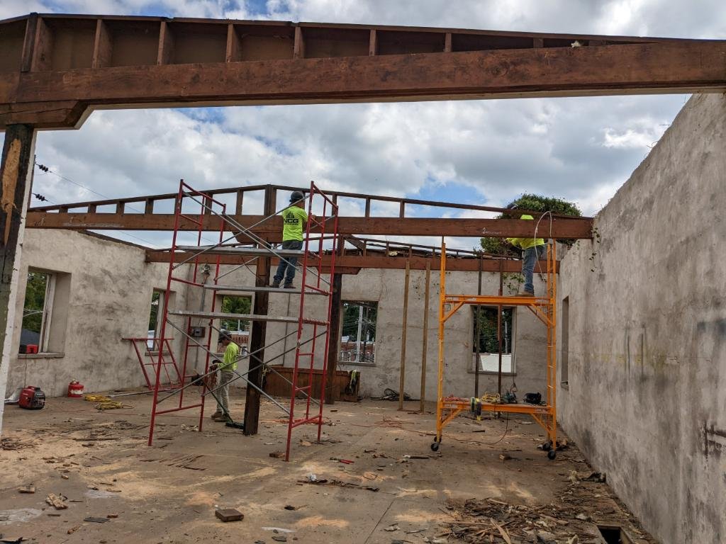 Three construction workers stand on scaffolding inside a partially demolished building, working on exposed steel beams under a mostly cloudy sky.