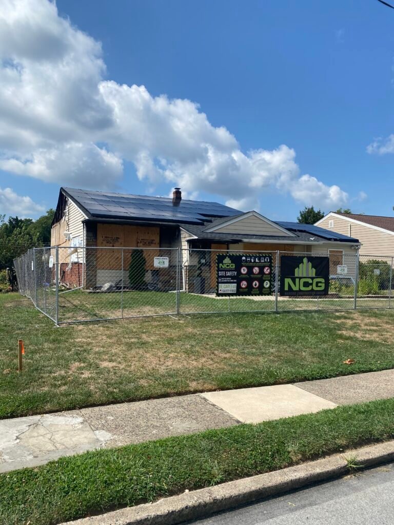 A house under renovation with boarded-up windows and doors, surrounded by a metal fence. A sign reading "NCG" and "Site Safety" is posted in front of the property.