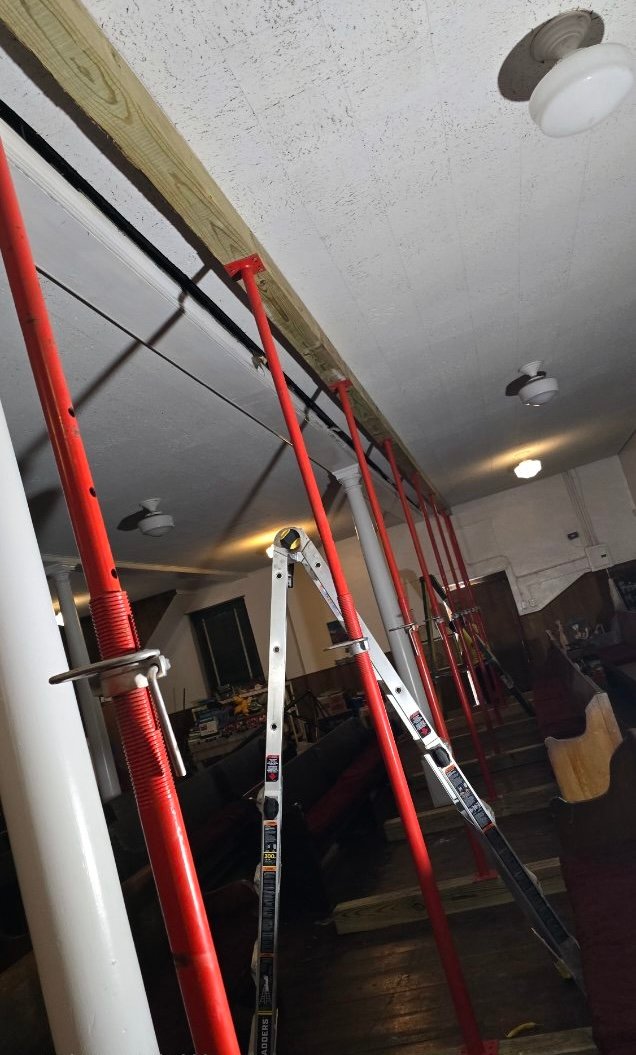 Multiple red support jacks and a ladder hold up a wooden beam under a white ceiling in a room with pews and scattered tools.