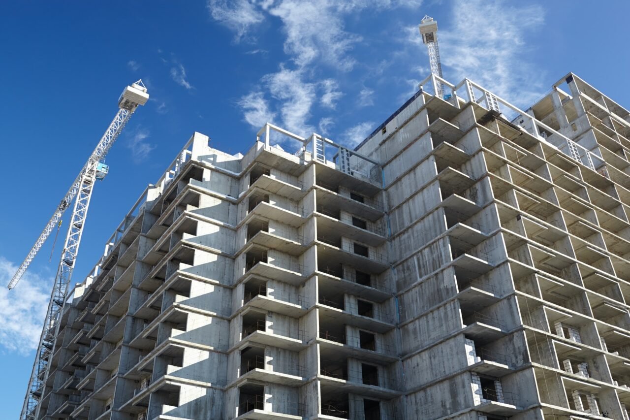Concrete frame of a multi-story building under construction with two cranes and a partly cloudy blue sky in the background.