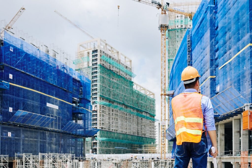 A construction worker wearing a safety vest and helmet observes several high-rise buildings under construction, surrounded by cranes and scaffolding.